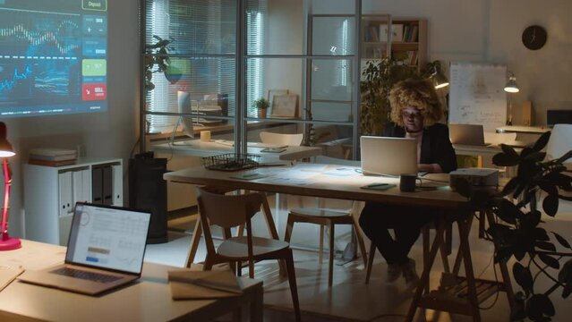 Wide shot of African American businesswoman using laptop at desk in dark office of investment company while overworking late at night