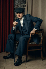 A young man with a cane is sitting in an armchair. English retro gangster of the 1920s dressed in a...