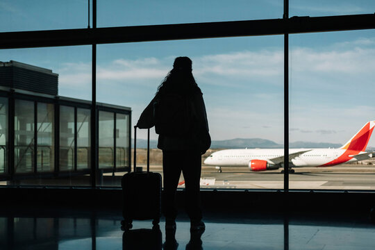 Silhouette of a woman with her luggage looking out the window at an airport. Travel concept.