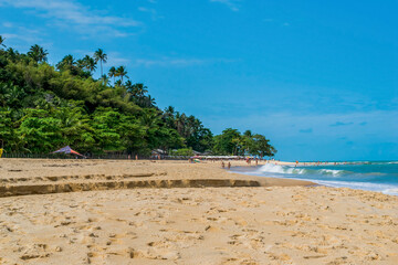 Linda praia com areias escuras, mar com ondas, em frente um pequeno rio chegando ao mar, pessoas passeando ao fundo e céu azul em Trancoso, Bahia.