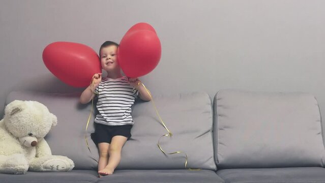baby boy kid in striped t-shirt jumping on grey sofa playing two red heart shape balloons. caucasian child having fun home coach rolling, lying, laughing. anniversary, birthday, Mother's Day holiday