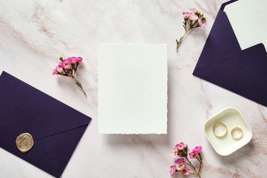 Wedding invitation card template, purple envelopes, rings, pink flowers on stone desk table. Flat lay, top view, copy space.