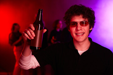 Portrait of young man holding beer bottle at party and smiling at camera lit by neon light