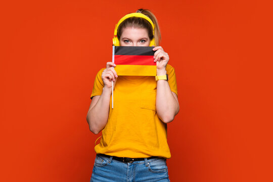 Learning German, student exchange and travel. Happy lady holding small flag of Germany, having fun. Girl peeking out from behind flag. Ready to study in college, standing over orange background