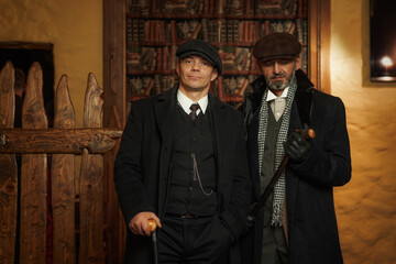 Two men of different ages, English retro gangster of the 1920s dressed in a coat, suit and flat cap in Peaky blinders style. - obrazy, fototapety, plakaty