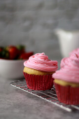 Close up of vanilla muffins with pink strawberry buttercream frosting on top, grey marble table and white stone wall in background