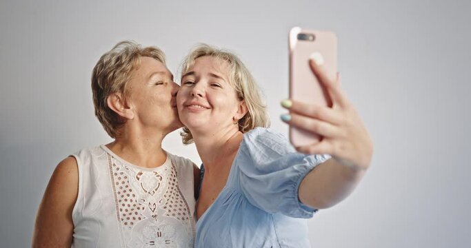 Mother and daughter kissing and taking selfie. Adult daughter smiling and kissing mature mother while taking selfie via cellphone together against gray background