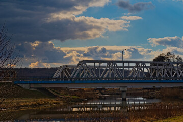 Dwa mosty - drogowy i kolejowy - rozpięte nad rzeką Kamienną  , na tle błękitnego nieba i chmur.
