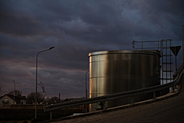 Przemysłowy wielki zbiornik ,  przeciwpożarowy na wodę ,  ze stali nierdzewnej na tle ciemnych chmur i oświetlony promieniami zachodzącego słońca .