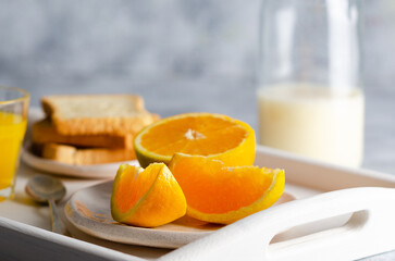 A bottle of milk, a glass of orange juice, and pieces of orange, in a white tray and grey background. 