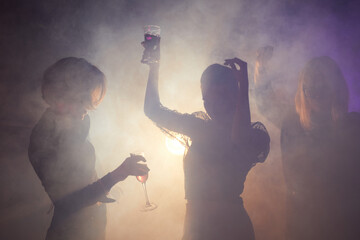 Backlit shot of young women dancing at party, elegant silhouettes in smoky club