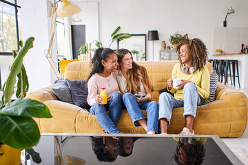 Three happy female friends smiling and having fun at home - Funny women together celebrating sitting on the living room sofa drinking juice and coffee