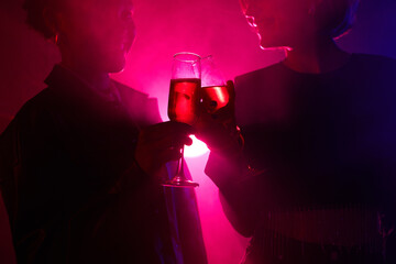 Backlit portrait of two young women enjoying drink while partying in smoky club with neon lights,...