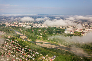 Aerial view of white clouds above a town or village with rows of buildings and curvy streets between green fields in summer. Countryside landscape from above.