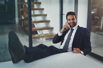 Business just went from good to great. Shot of a handsome young businessman relaxing with his feet up on an office desk while using his phone.