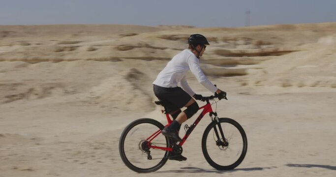 Tracking shot of male biker riding red mountain bike in desert dirt track, Manama Bahrain, day