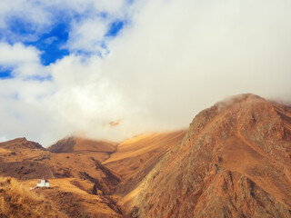 Golden Caucasian mountain ranges with clouds descending on them and with a white house located on...