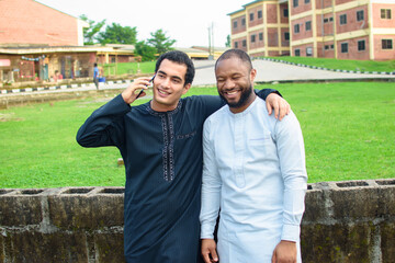 Two fair complexion african male friends or students happily standing together in an outdoor environment while one of them is making call with a smart android phone