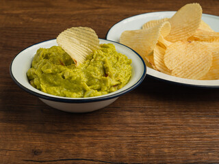 A bowl with fresh guacamole and potatoes chips on a white plate over a wooden table
