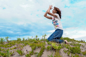Fototapeta premium Adult latin woman smiling and doing yoga in the beach receiving the spring season in a praying pose and close eyes, feeling the fresh air. Freedom and joyful lifestyle concept.