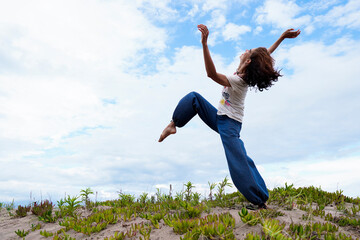 Adult latin woman in the dunes celebrating springtime season with a dance movement while looking up the clouds in the sky. Breathing fresh air. Freedom and joyful lifestyle concept.  
