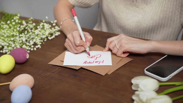 Close-up on a table with items to create a composition for Easter. A woman's hand writes with a red marker: Happy Easter. Christ is risen. Church holiday-Easter