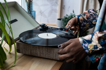 Hands of young black man with nail polish putting vinyl record on needle player standing on wooden windowsill before listening retro music
