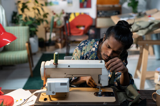 Young creative male tailor sitting by workplace in front of sewing machine while putting thread through needle eyes
