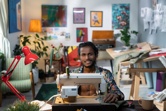 Young successful gender-neutral male designer of fashionable apparel working by sewing machine while sitting in workshop - Powered by Adobe
