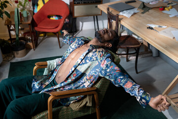 Young relaxed non-binary man in stylish clothes lying in armchair with his arms outstretched while...