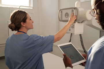 Young female clinician in uniform pushing button on panel of x-ray machine while preparing for examination of patients in hospital