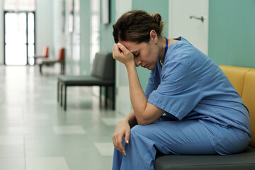 Young tired or stressed female doctor in uniform sitting in hospital corridor with her head in...