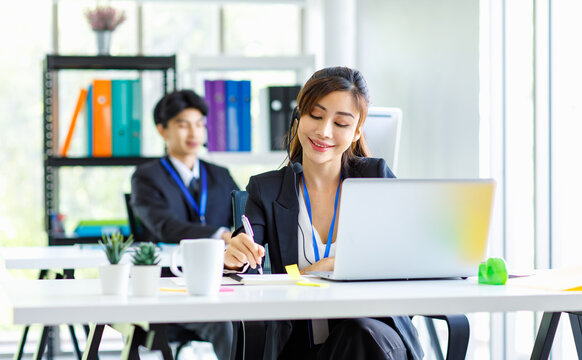 Asian young beautiful female businesswoman operator wearing headset with microphone working at helpdesk service with male colleague via laptop computer helping consulting with customers call center