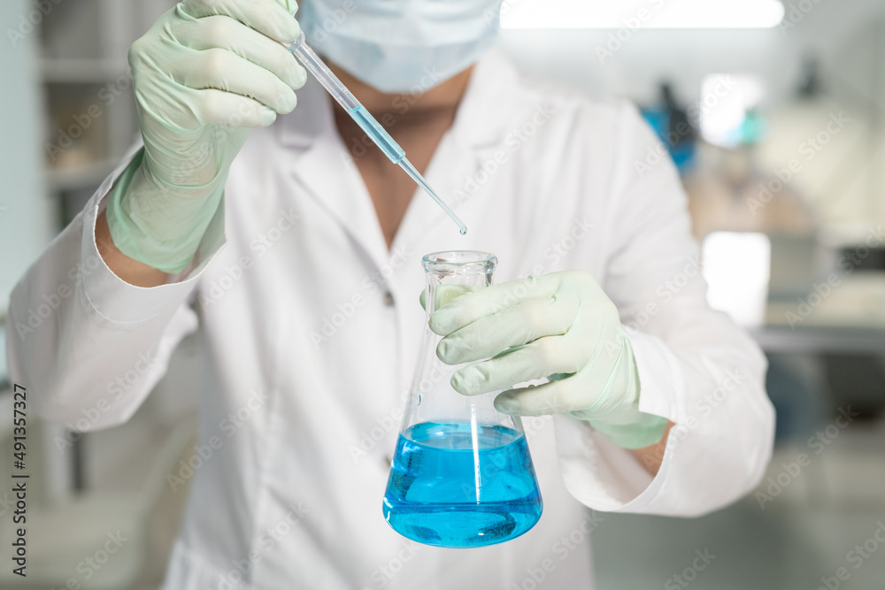 Aufkleber Contemporary scientist in surgical gloves, mask and lab coat adding drop of liquid into flask with blue chemical substance