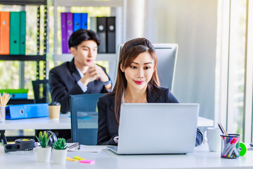 Asian young female professional businesswoman secretary employee in formal suit sitting at working desk typing report via laptop notebook computer while businessman manager work behind in office