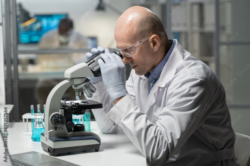 Poster experienced chemist in lab coat and surgical gloves studying new chemical element in microscope in s