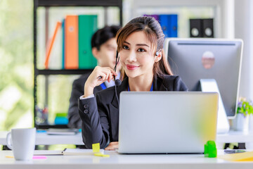 Asian young beautiful female businesswoman operator wearing headset with microphone working at helpdesk service with male colleague via laptop computer helping consulting with customers call center