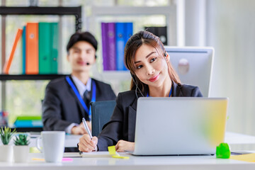 Asian young beautiful female businesswoman operator wearing headset with microphone working at helpdesk service with male colleague via laptop computer helping consulting with customers call center