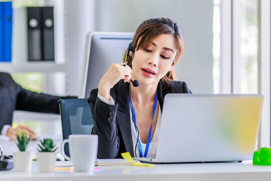 Asian young beautiful female businesswoman operator wearing headset with microphone working at helpdesk service with male colleague via laptop computer helping consulting with customers call center.