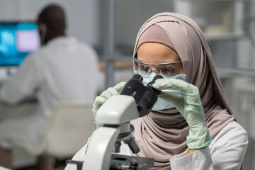 Contemporary female researcher in hijab looking in microscope while studying new chemical...