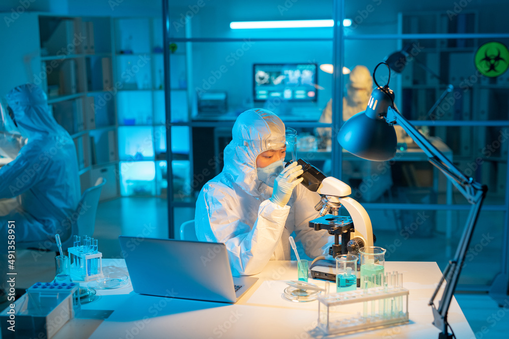 Poster female scientist in biohazard suit, respirator and gloves using microscope for studying new substanc