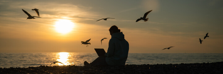 Caucasian woman typing on a laptop on the seashore at sunset. Freelance work. Wide screen. 