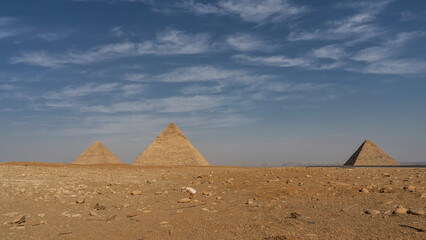 Fototapeta na wymiar Three Great Pyramids Giza against blue sky and clouds. Stones are scattered on the sand of the desert. Silhouettes of modern buildings of Cairo are visible on the horizon. Egypt