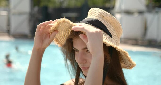 Beautiful young brunnete girl in straw hat smiling at camera near pool