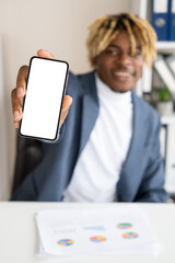 Business communication. Happy black man. Mobile application. Smiling defocused handsome guy showing smartphone blank screen sitting desk in light room interior.