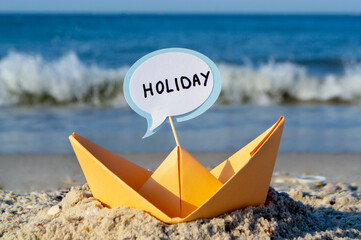 Orange Paper Boat on Sandy Seashore Close-up. Small Boat from Paper on Background of Blue Sea on Sunny Summer day. Stick with Word Holiday Inserted into Boat. Concept Travel, Tourism, Vacation, Rest