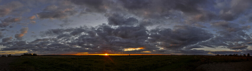 Wide panorama of flat landscape with dark clouds at sunset and orange sun