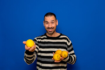 Hispanic man with a beard dressed in a striped sweater offering some lemons that he is holding in both hands isolated on blue studio background.
