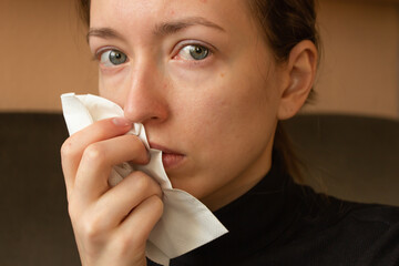 Portrait of young ill woman with runny nose sneezes into a napkin. Sick day at home. Illness cold...