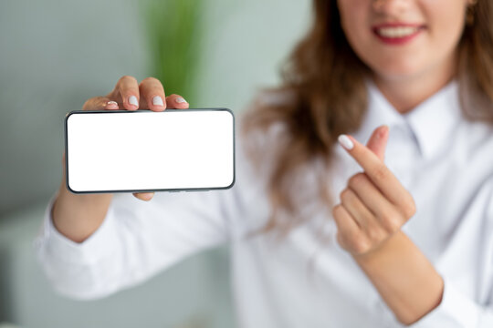 Small heart sign. Office woman. Mobile technology. Pretty unrecognizable smiling lady showing smartphone blank screen with love finger gesture in light room interior defocused.